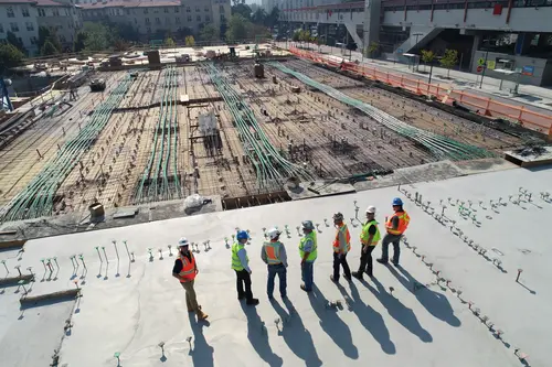 Sieben Männer in Sicherheitskleidung überblicken eine große, weitläufige Baustelle unter freiem Himmel.